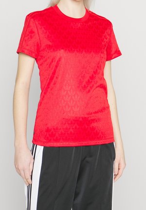 Czerwona sportowa koszulka z krótkim rękawem z subtelnymi tłoczonymi wzorami, noszona z czarno-białymi spodniami w paski, pokazująca górną część tułowia i ramiona.
