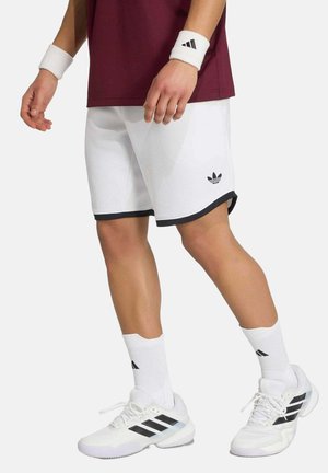 Persoon draagt witte Adidas shorts, witte sokken, witte Adidas sneakers en een kastanjebruin shirt met witte polsbanden tegen een effen achtergrond.