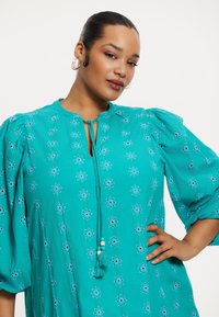Türkise Bluse mit Puffärmeln, ausgestattet mit floralem Stickerei und einem Schlüssellochausschnitt, der mit einem dekorativen Band und Quastenverzierungen gebunden ist.