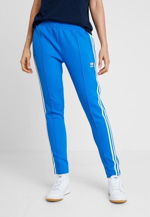 Persoon draagt felblauwe Adidas trainingsbroek met witte zijstrepen en witte sneakers, staand tegen een effen achtergrond.