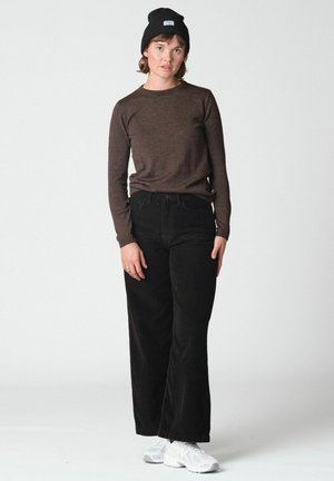 Jeune femme portant une chemise marron à manches longues, un pantalon noir à jambes larges, des baskets blanches et un bonnet noir, debout devant un fond uni.