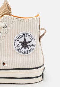 Sapatilha de lona com parte superior em bege canelado, retalho branco com uma estrela e texto, forro laranja e sola de borracha preta.