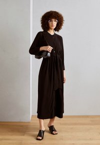 Abito nero con bottoni, vita legata, maniche lunghe e silhouette fluida. Abbinato a sandali in pelle nera open-toe.
