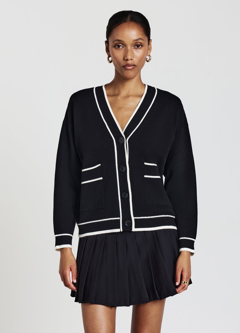 Cardigan noir avec des bordures blanches, décolleté en V et poches avant. Il comporte cinq boutons noirs et une coupe décontractée, associé à une jupe plissée noire.
