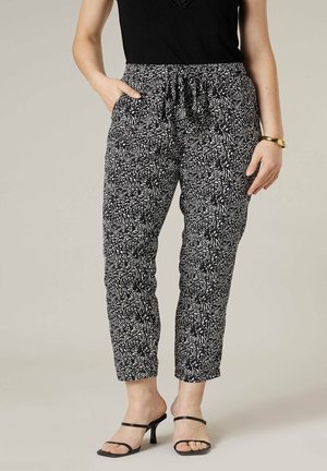 Femme portant un pantalon à cordon noir et blanc à motifs, un haut noir sans manches, des sandales à talons noires et un bracelet en or.