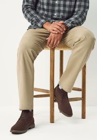Hnedé semišové členkové topánky so šnurovaním, nosené s béžovými nohavicami. Jednotlivec sedí na drevenej stoličke, ruky zložene na kolenách.