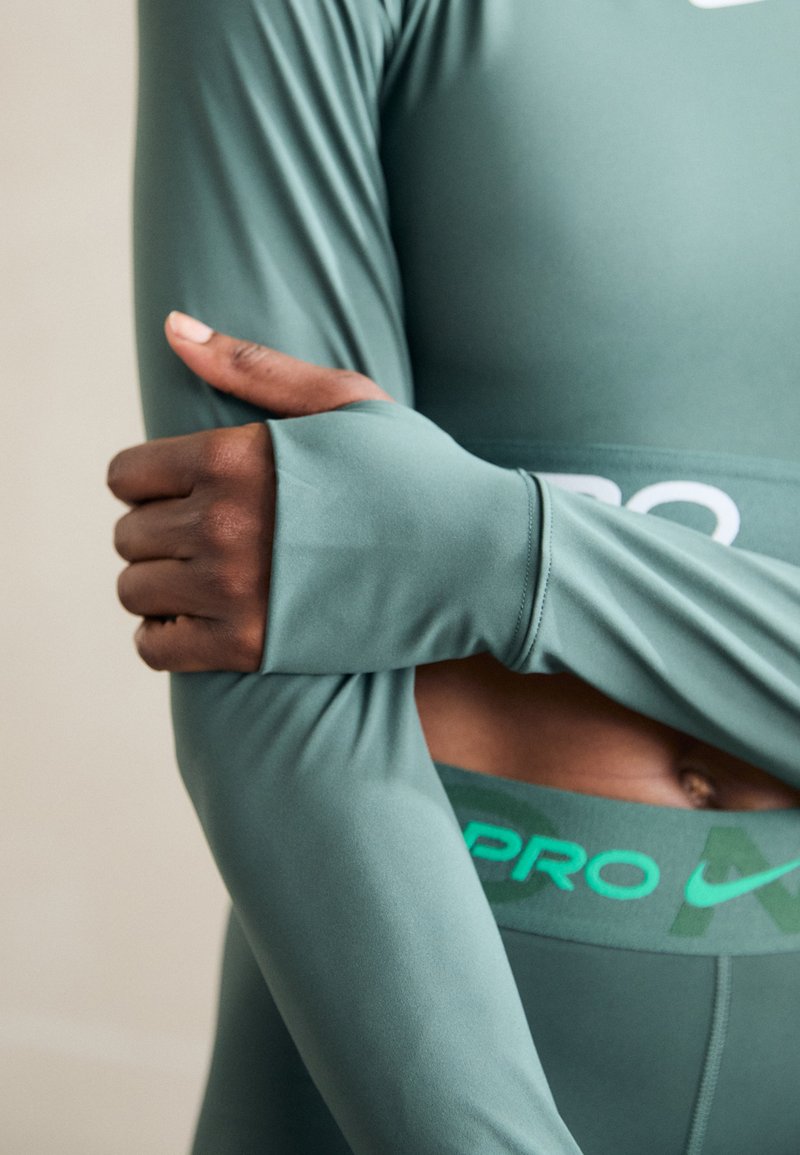 Maglietta athletic a maniche lunghe in verde tenue, con una texture elegante e aperture per i pollici. Una fascia con logo verde mette in risalto i leggings a vita alta sottostanti.
