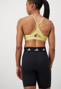 Sujetador deportivo amarillo con un diseño de patrón y un corte en la espalda, combinado con pantalones cortos ajustados negros que presentan un logo blanco en la cintura.