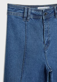 Denim hlače v srednje modri barvi z gladko teksturo, ki vključujejo klasičen dizajn s petimi žepi in standardnim gumbom za zapiranje.
