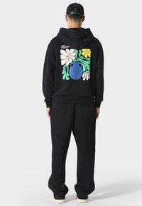 Musta huppari ja collegehousut -setti, jossa on värikäs kukkakuosi ja sininen vaasi takana. Kangas vaikuttaa pehmeältä ja mukavalta.