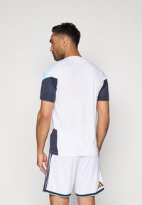 Λευκή αθλητική φανέλα με ναυτικές και ανοιχτό μπλε λεπτομέρειες, κοντά μανίκια και v-neck. Συνδυάζεται με λευκές σορτς που διαθέτουν σκούρες ρίγες και λογότυπο.