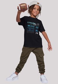 Black t-shirt featuring blue car illustrations and text. Paired with olive cargo pants and white sneakers. Holding a brown football.
