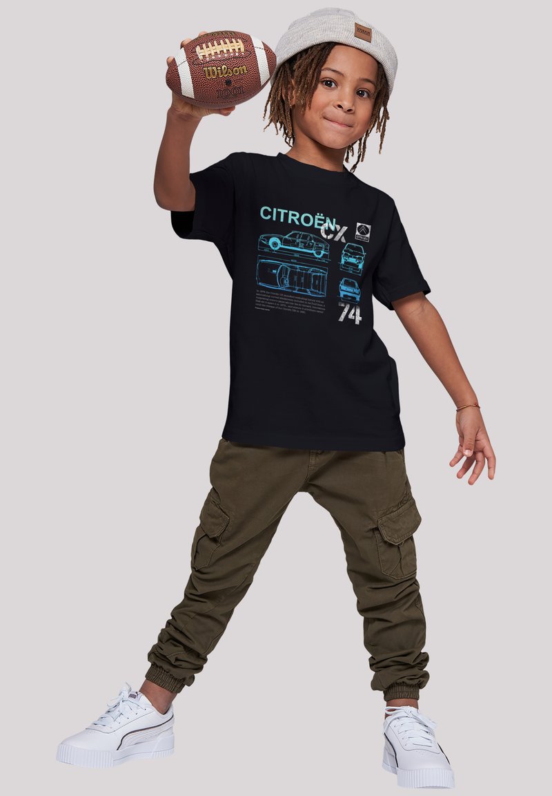 Black t-shirt featuring blue car illustrations and text. Paired with olive cargo pants and white sneakers. Holding a brown football.
