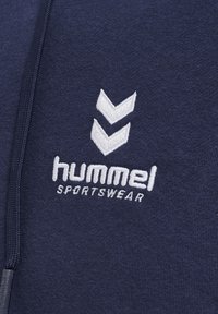 Sweat à capuche bleu marine doté d'une fermeture éclair, d'une texture douce et du logo blanc "hummel" brodé avec deux chevrons au-dessus.