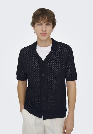 Giovane uomo che indossa una camicia nera a maniche corte con bottoni sopra una T-shirt bianca e pantaloni beige chiaro, in piedi su uno sfondo semplice.