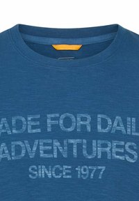 Blauw shirt met ronde hals en vervaagde tekst "MADE FOR DAILY ADVENTURES SINCE 1977" op de voorkant gedrukt.