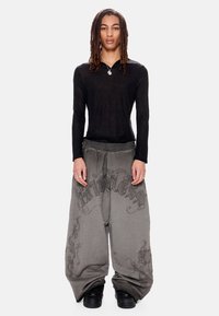 Νεαρός με μακριά dreadlocks φοράει διάφανο μαύρο τοπ με μακριά μανίκια, γκρι φαρδιά παντελόνια με σκοτεινά σχέδια και μαύρα αθλητικά παπούτσια.