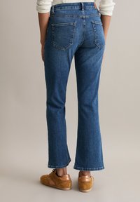 Woman wearing blue straight-leg jeans and brown suede sneakers, standing on a neutral background facing away from the camera.