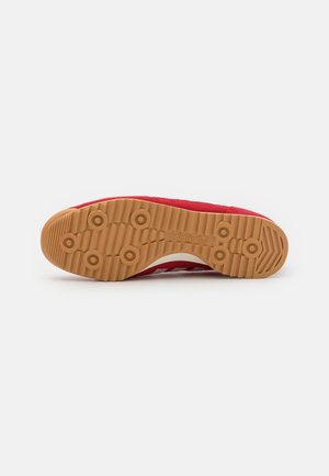 Czerwona podeszwa sneakersa z brązowym gumowym bieżnikiem z okrągłymi wypustkami i logo adidas na środku, sfotografowana na białym tle.