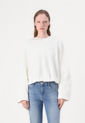 Witte longsleeve shirt met ronde hals, voorzien van verlaagde schouders en een zachte textuur. Draagt met blauwe jeans en een gouden knoopaccent.