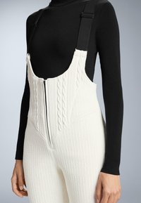 Fehér, kötött mintás kantáros nadrág elöl cipzárral és állítható fekete vállpántokkal, fekete hosszú ujjú felsővel párosítva. Texturált anyag.