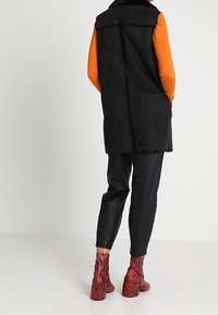 Svart konstpälsväst, orange långärmad tröja, svarta läderbyxor och röda ankle boots med ormtryck. Bakvy, jämna texturer, lagerdesign.