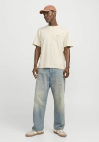 Beige katoenen t-shirt met een logo, gecombineerd met lichtblauwe recht gesneden denim jeans. G gedragen met een lichtbruine pet en casual sneakers.