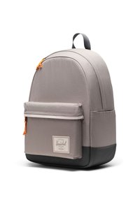 Sac à dos en tissu gris à la forme arrondie, avec un fond noir, une poche avant et un tirette de fermeture éclair orange. Présente un patch logo sur le devant.