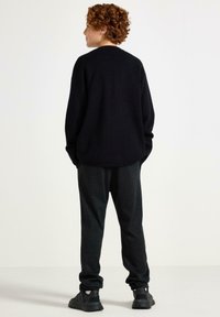 Czarny oversize sweter z długimi rękawami, zestawiony z ciemnozielonymi spodniami w kratę i czarnymi sneakersami. Widoczne kręcone włosy z tyłu.