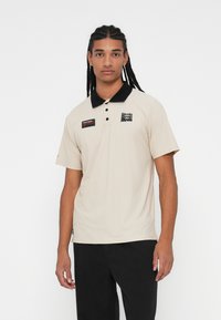 Beige polo shirt with a black collar, short sleeves, and a logo on the left chest. Features a tag and durable fabric.