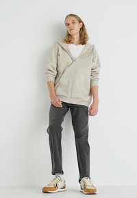 Veste à capuche zippée beige avec une capuche spacieuse et des poches avant, associée à un pantalon gris foncé et des baskets multicolores. Texture de tissu lisse.