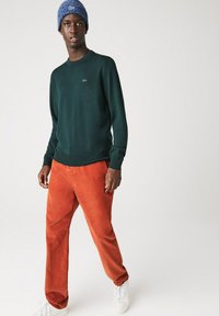 Maglione verde a maglia, pantaloni in corduroy arancioni, sneakers bianche e un berretto blu. Concentrati sulla texture e sul contrasto di colori nell'abbigliamento casual.
