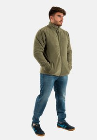 Giacca in pile verde oliva con zip completa, colletto alto e tasche laterali, abbinata a jeans in denim blu e sneakers nere con dettagli blu.