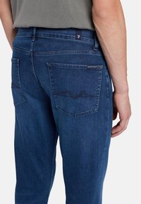 Jeans in denim blu scuro con un design aderente, caratterizzati da due tasche posteriori con cucitureCurve e un'etichetta con il marchio vicino alla vita.