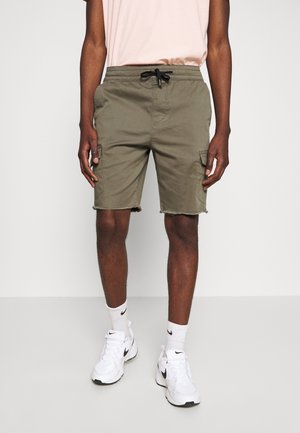 Mann in olivgrünen Cargo-Shorts mit ausgefransten Säumen, einem hellrosa Hemd, weißen Nike-Socken und weißen Nike-Sneakern, der an einer schlichten Wand steht.