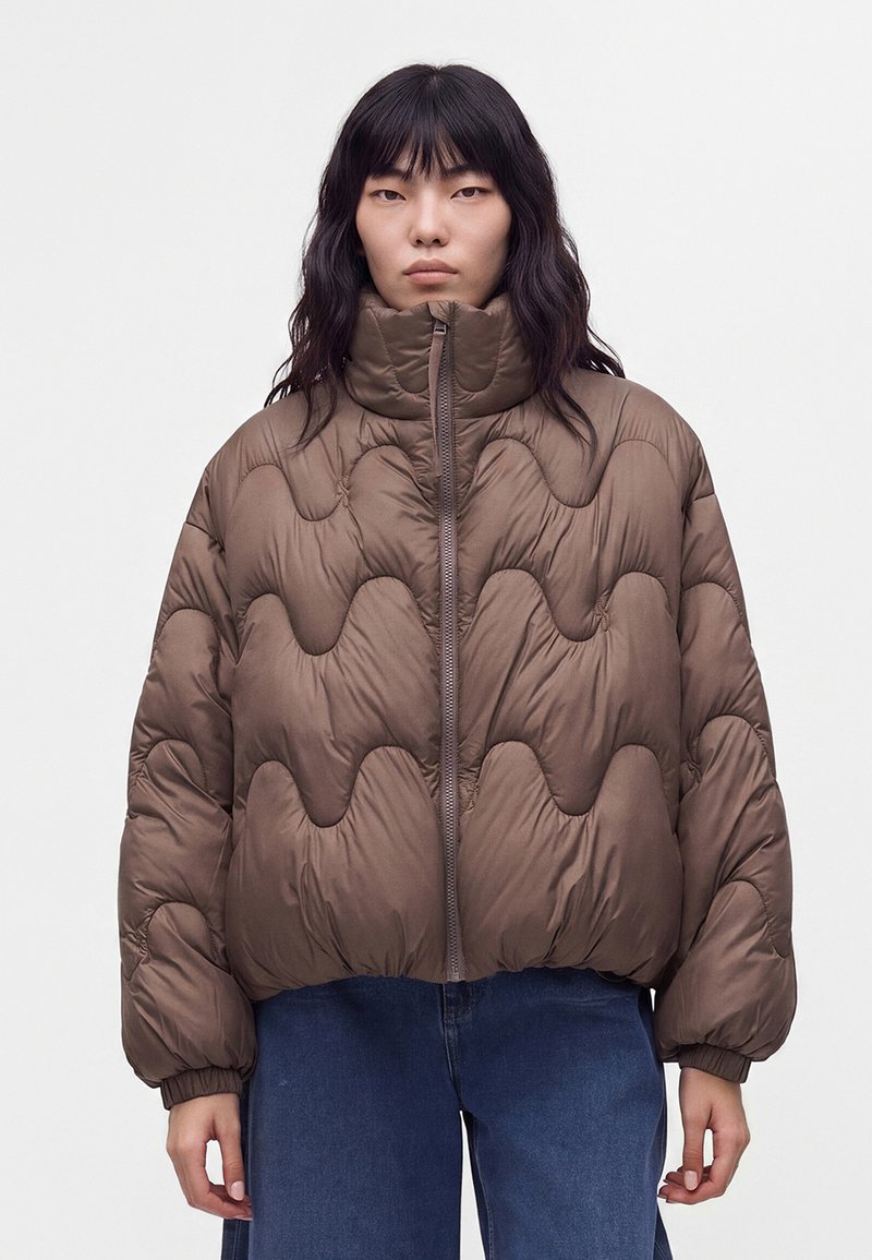 Femme aux longs cheveux foncés portant une doudoune matelassée marron et un jean bleu sur un fond clair uni.