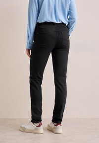 Jeans negros con un diseño ajustado, bolsillos traseros y detalles de costura, combinados con zapatillas blancas y marrón burdeos, vistos desde la parte posterior.