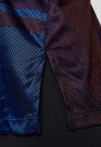 Tessuto testurizzato con un motivo blu e bordeaux, dotato di uno spacco laterale. Il materiale appare traspirante e aderente su uno sfondo nero solido.
