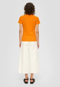 T-shirt en coton orange à manches courtes, associé à un pantalon en denim blanc à jambes larges. Des sandales texturées complètent l'ensemble.