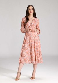Midi dress floral berwarna pink lembut dengan aksen oranye dan ungu. Memiliki leher V, lengan panjang, dan rok mengalir. Model mengenakan sepatu hak emas.