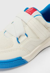 White athletic shoe with blue accents, textured mesh, and hook-and-loop straps. Features a rubber sole and a red label on the heel.