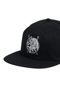 Black cap made of fabric featuring a circular white graphic with intertwined designs, including a creature and chains, positioned on the front.