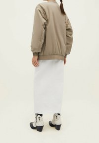 Lichtbeige bomberjack met elastische zoom, gecombineerd met een lange witte denimrok en crème enkellaarsjes met zwarte accenten.