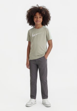 Göndör hajú gyermek világoszöld Nike pólóban, sötétszürke nadrágban és fehér tornacipőben, egy kézzel a zsebében áll.