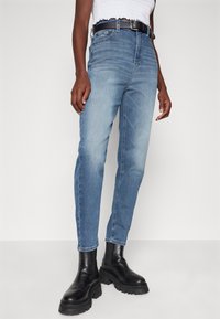 Modré džíny s rovného střihu, s vybledlým vzhledem, pěti kapsami a černým opaskem, nošené s masivními černými botami.