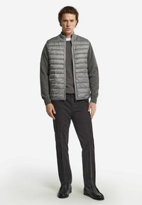 Grau gepolsterte Weste über einem dunkelgrauen Pullover, kombiniert mit schwarzen Hosen und Schuhen. Die Weste hat ein vertikales Steppmuster und einen hohen Kragen.