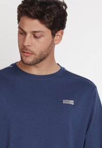 Felpa girocollo blu navy realizzata in cotone, caratterizzata da un piccolo etichetta con testo e un'accentuazione cucita sulla spalla.