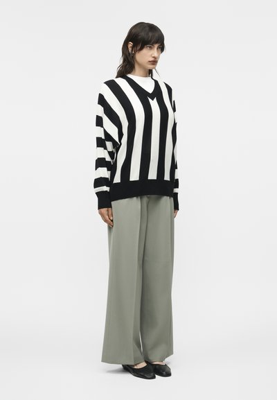 Γυναίκα που στέκεται φορώντας πουλόβερ με κάθετες ασπρόμαυρες ρίγες, φαρδιά παντελόνια ανοιχτό γκρι και μαύρα μυτερά παπούτσια σε λευκό φόντο.