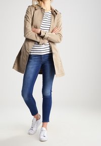Beige trenchcoat met een knoopsluiting, gedragen over een wit en marineblauw gestreept shirt. Gepaard met blauwe skinny jeans en witte sneakers.