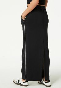 Person iført en sort maxi-nederdel med en hvid sidestribe og sideslids, kombineret med hvide og sorte sneakers, med hånden i lommen.